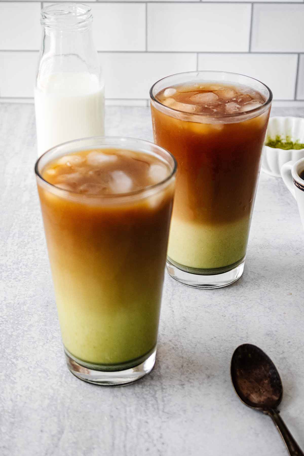 Two iced matcha coffee drinks in tall glasses with a spoon and bowl of matcha powder