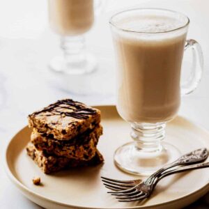 Two chai tea lattes in glass mugs. The front latte is sitting on a creamy white plate with two forks and a stack of granola bars.