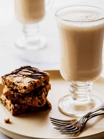 Two chai tea lattes in glass mugs. The front latte is sitting on a creamy white plate with two forks and a stack of granola bars.
