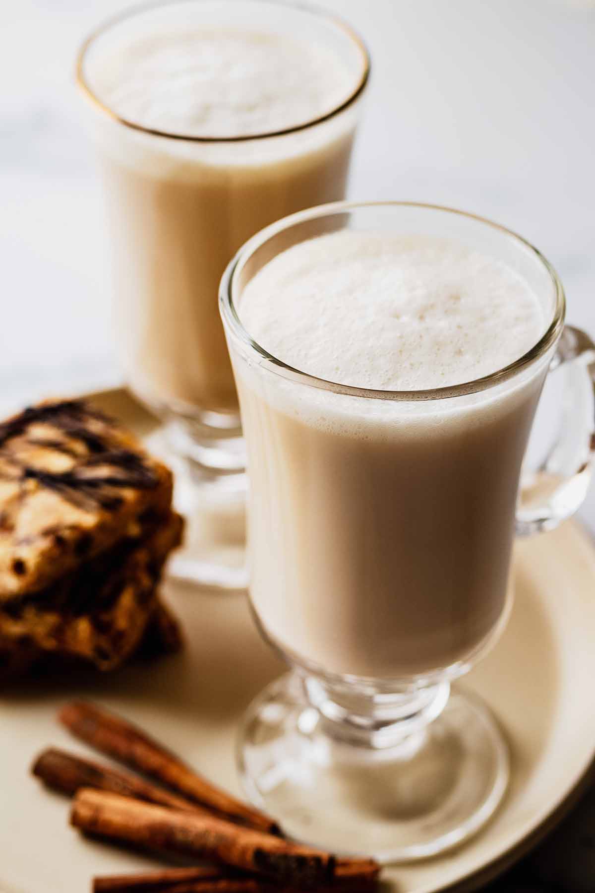 Close up view of two chai tea lattes in glass mugs sitting on a creamy white plate with a stack of granola bars and cinnamon sticks.