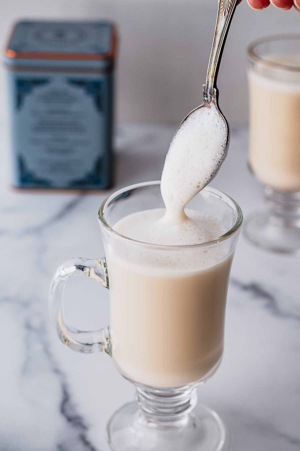 Latte in a tall glass mug with foam being added to the top with a spoon.