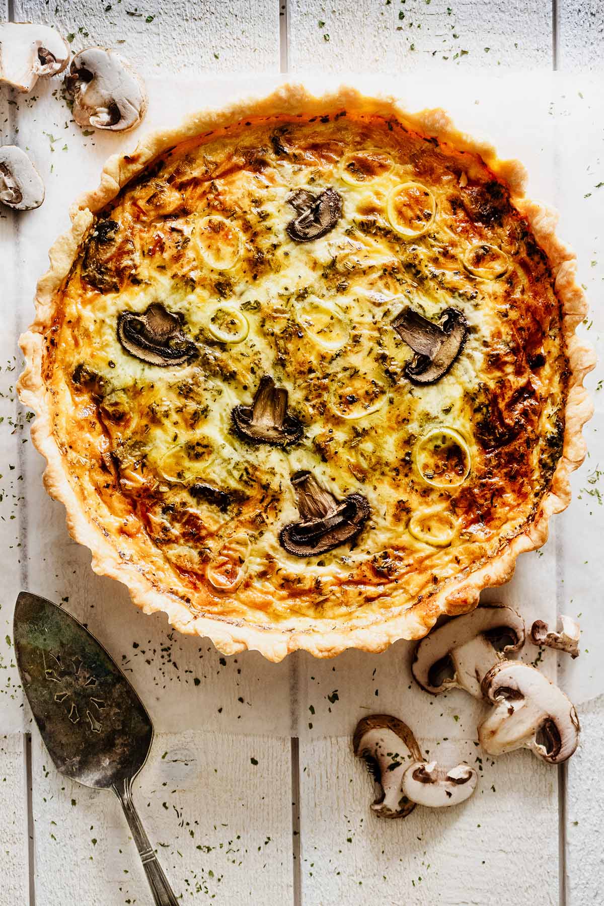 Overhead view of whole quiche on a white wood background with mushrooms and a serving spatula
