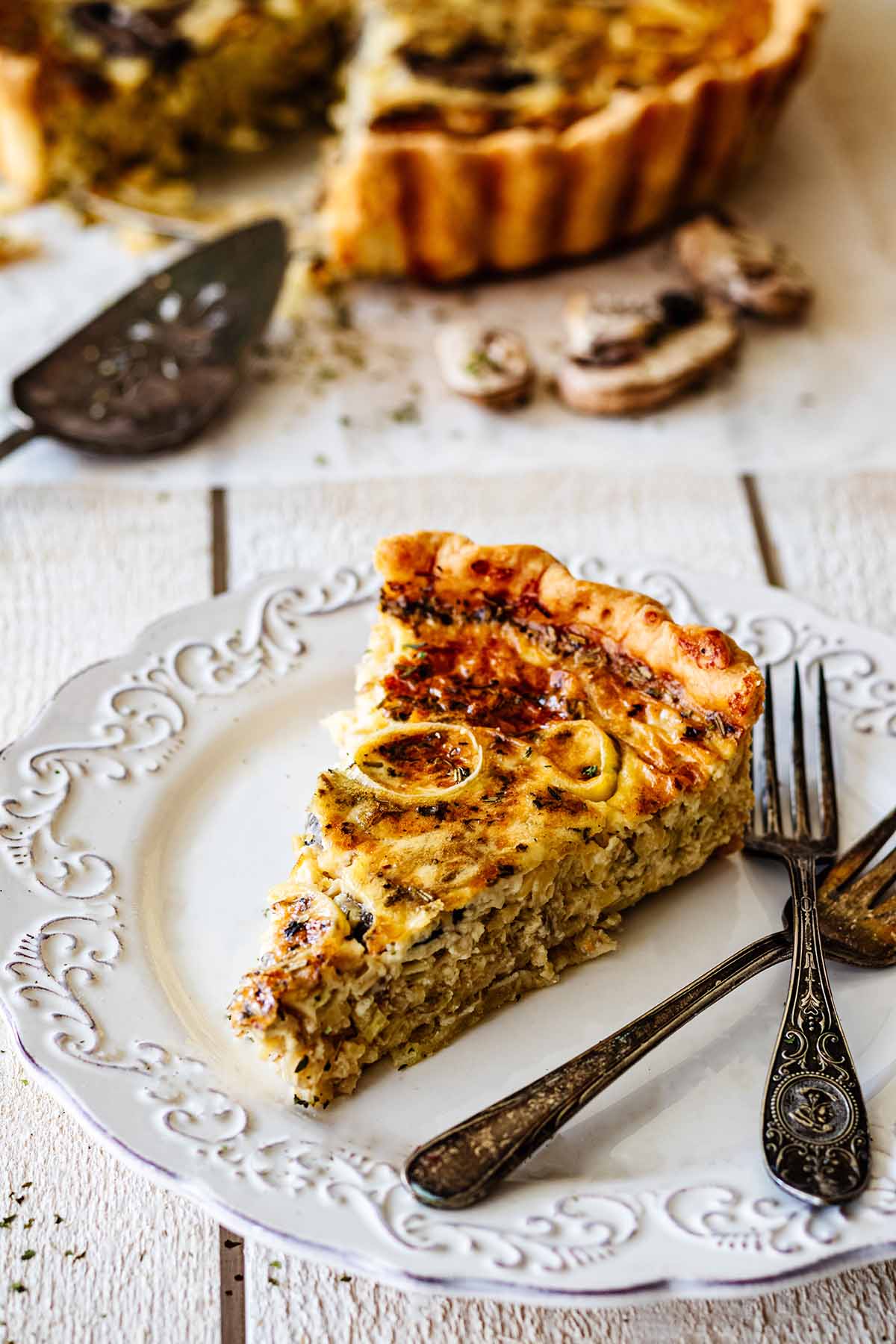 Slice of leek and mushroom quiche on a white plate with two forks. A whole quiche with a slice taken out is in the background.