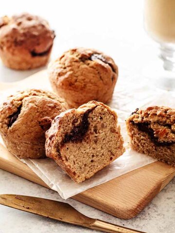 Four Nutella muffins on a small cutting board. One of the muffins is cut in half to reveal the Nutella filling. A gold knife and glass mug filled with tea are off to the side.