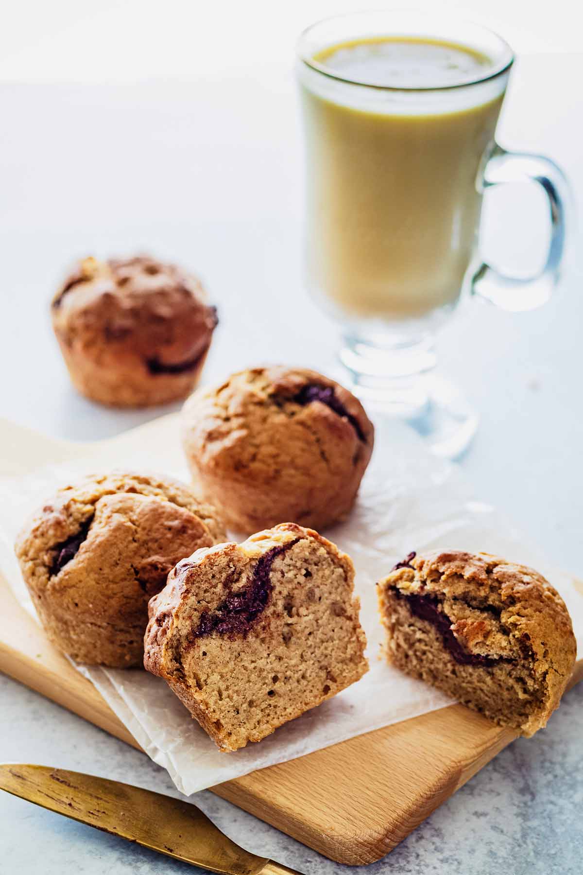 Four Nutella muffins on a small cutting board. One of the muffins is cut in half to reveal the Nutella filling. A gold knife and glass mug filled with tea are off to the side.
