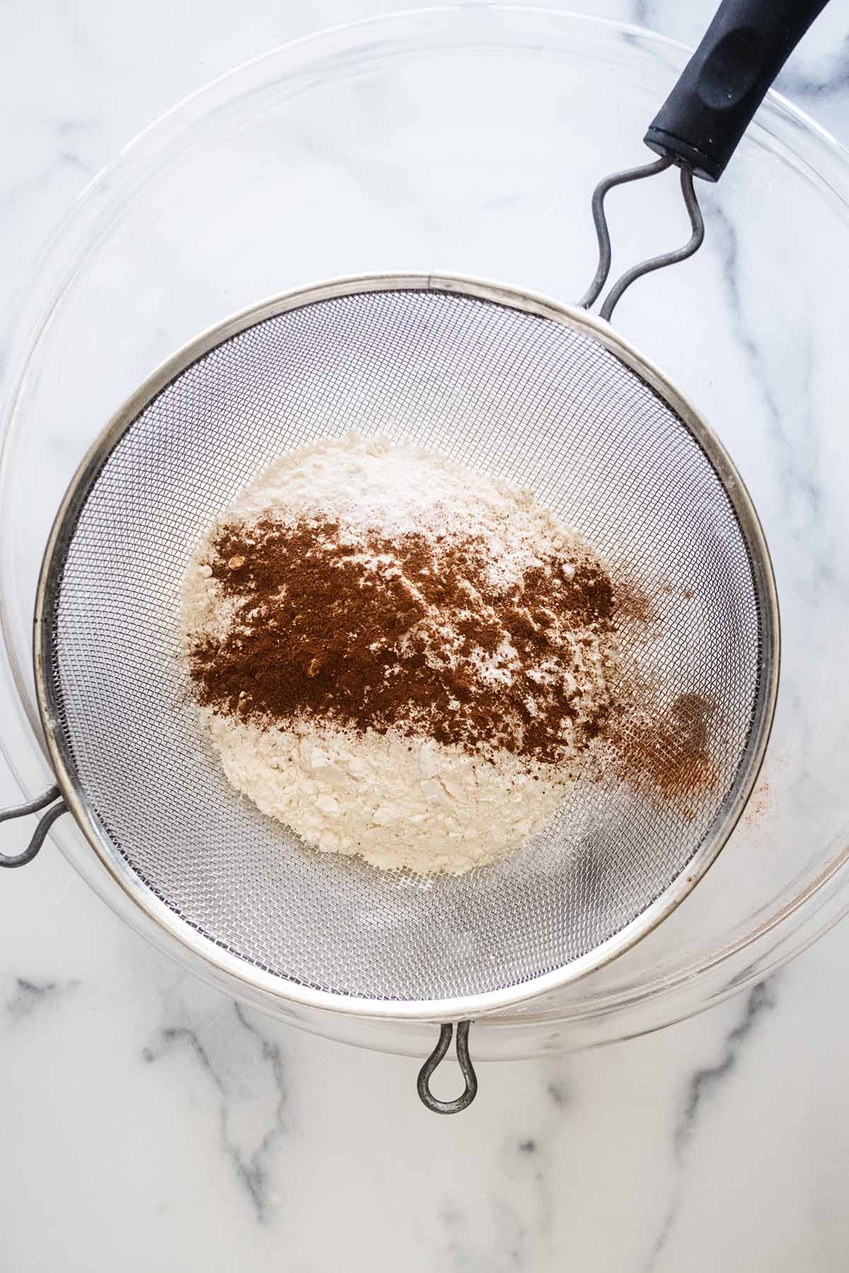 Dry ingredients in a mesh strainer over a large glass bowl.