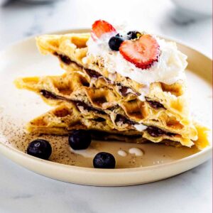 Stack of Nutella waffles cut on a diagonal and topped with whipped cream and fresh berries on a creamy white plate