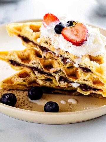 Stack of Nutella waffles cut on a diagonal and topped with whipped cream and fresh berries on a creamy white plate