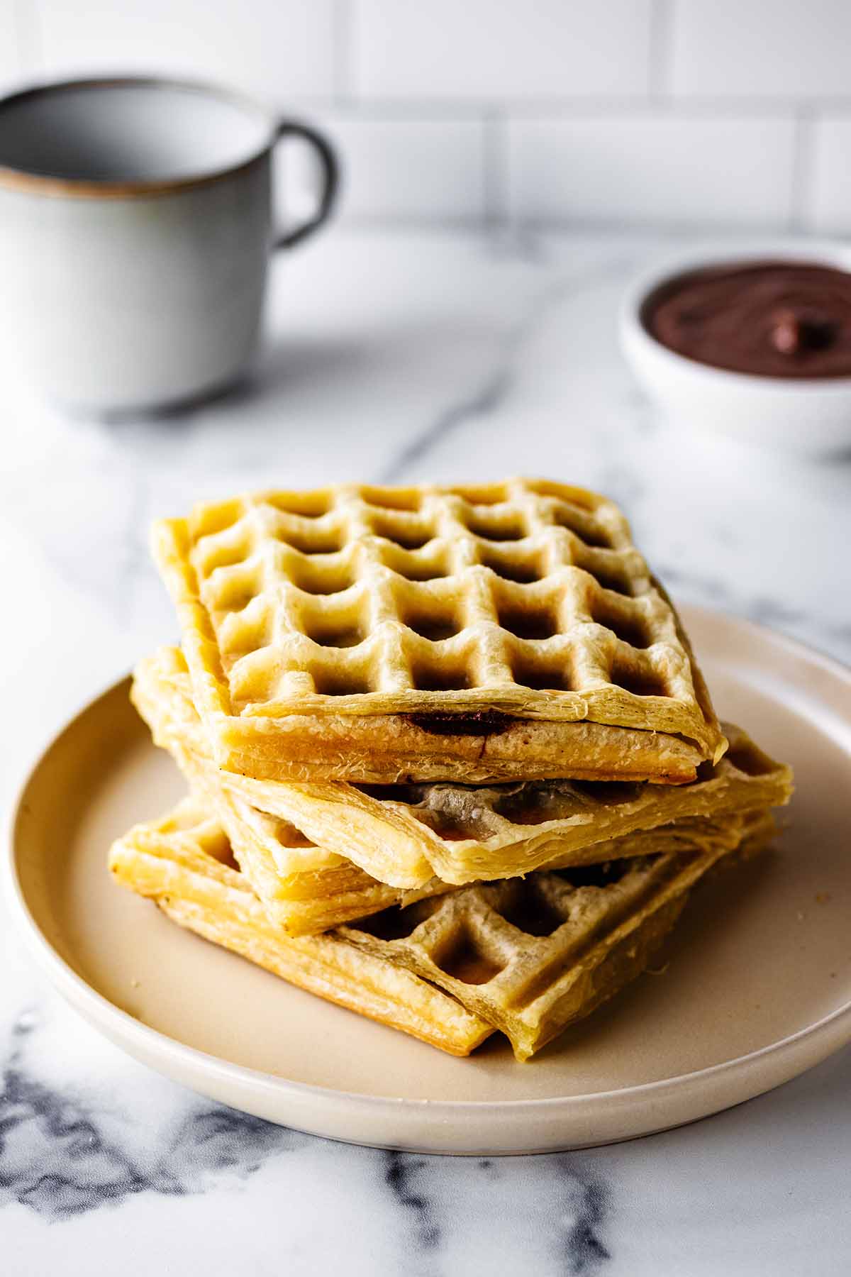 Stack of Nutella waffles on a creamy white plate a bowl of Nutella and coffee mug are in the background