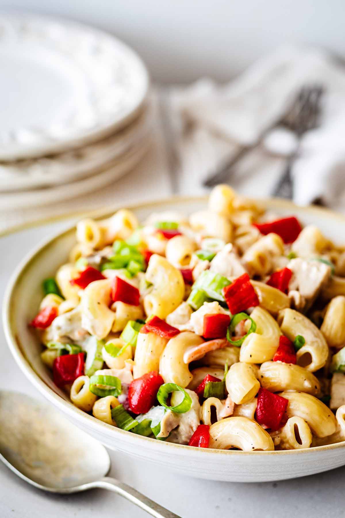 Closeup of chicken macaroni salad in a white bowl with a metal serving spoon and a stack of white plates in the background.