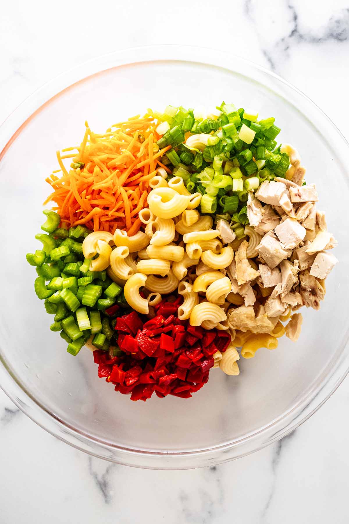 Overhead view of cooked macaroni, chopped vegetables and dressing in a large glass bowl.