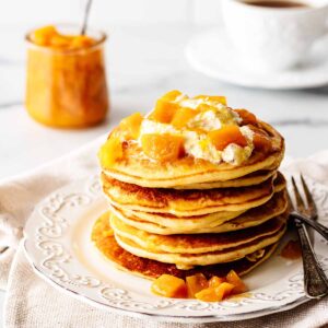 Stack of mango pancakes topped with whipped cream and mango compote on a white plate with a fork.