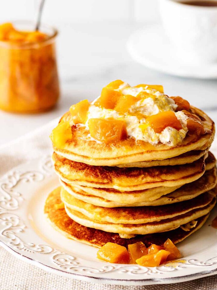 Stack of mango pancakes topped with whipped cream and mango compote on a white plate with a fork.