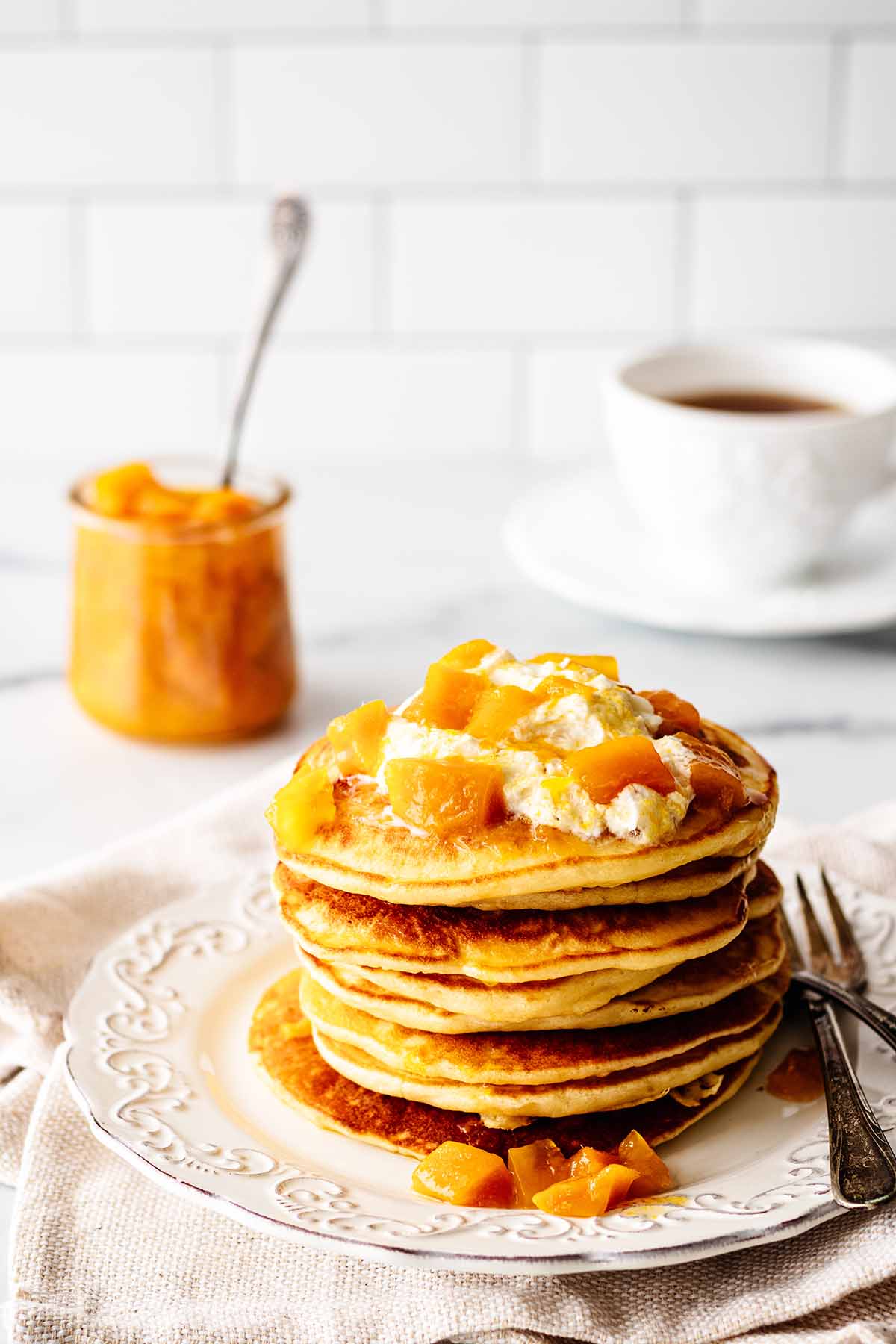 Stack of pancakes topped with whipped cream and mango compote on a white plate with two forks. A jar of fresh mango compote and hot tea in a white teacup with saucer are in the background.