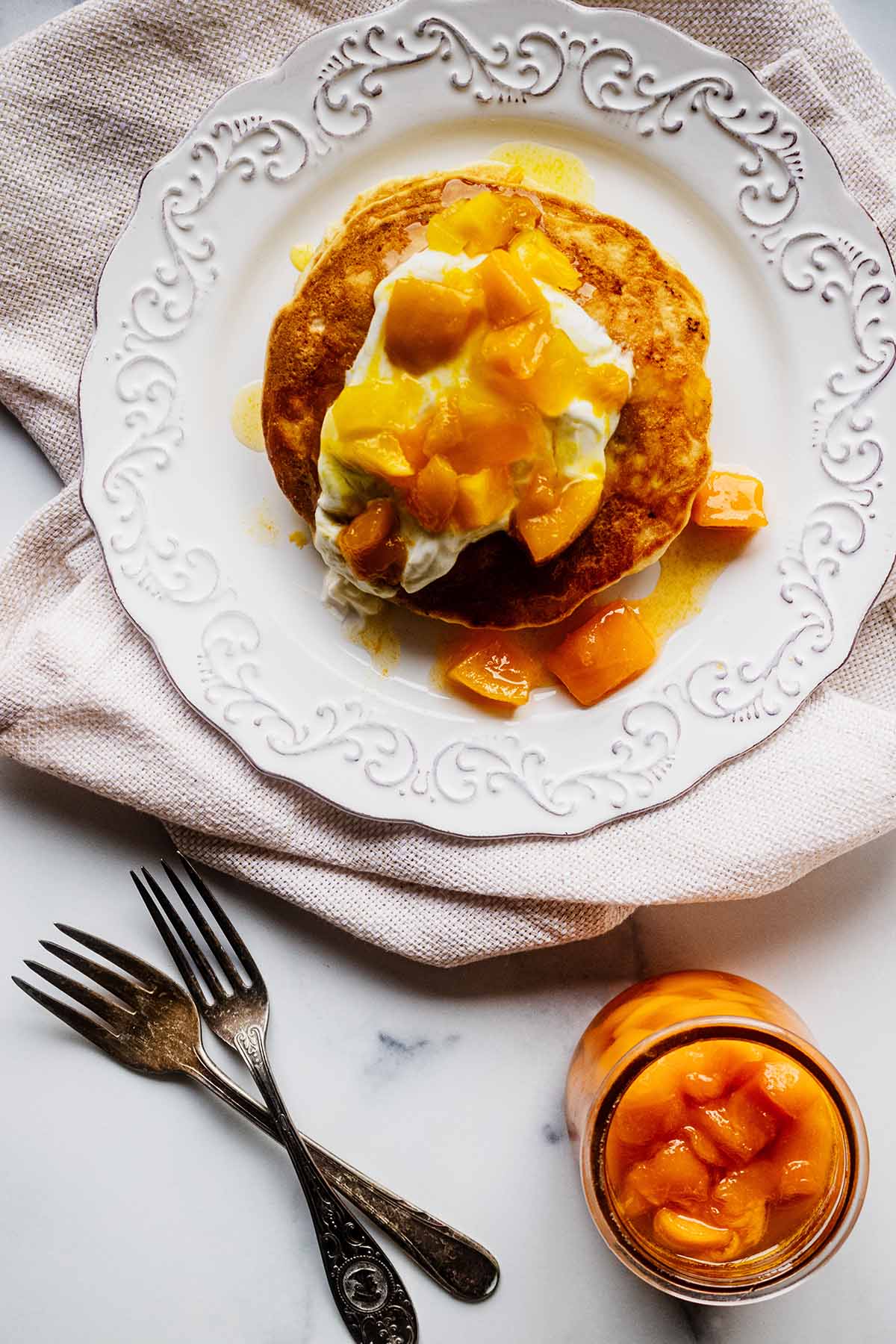 Overhead view of mango pancakes topped with yogurt and mango compote on a white plate on a muslin napkin. Two forks and a glass jar of mango compote are off to the side.