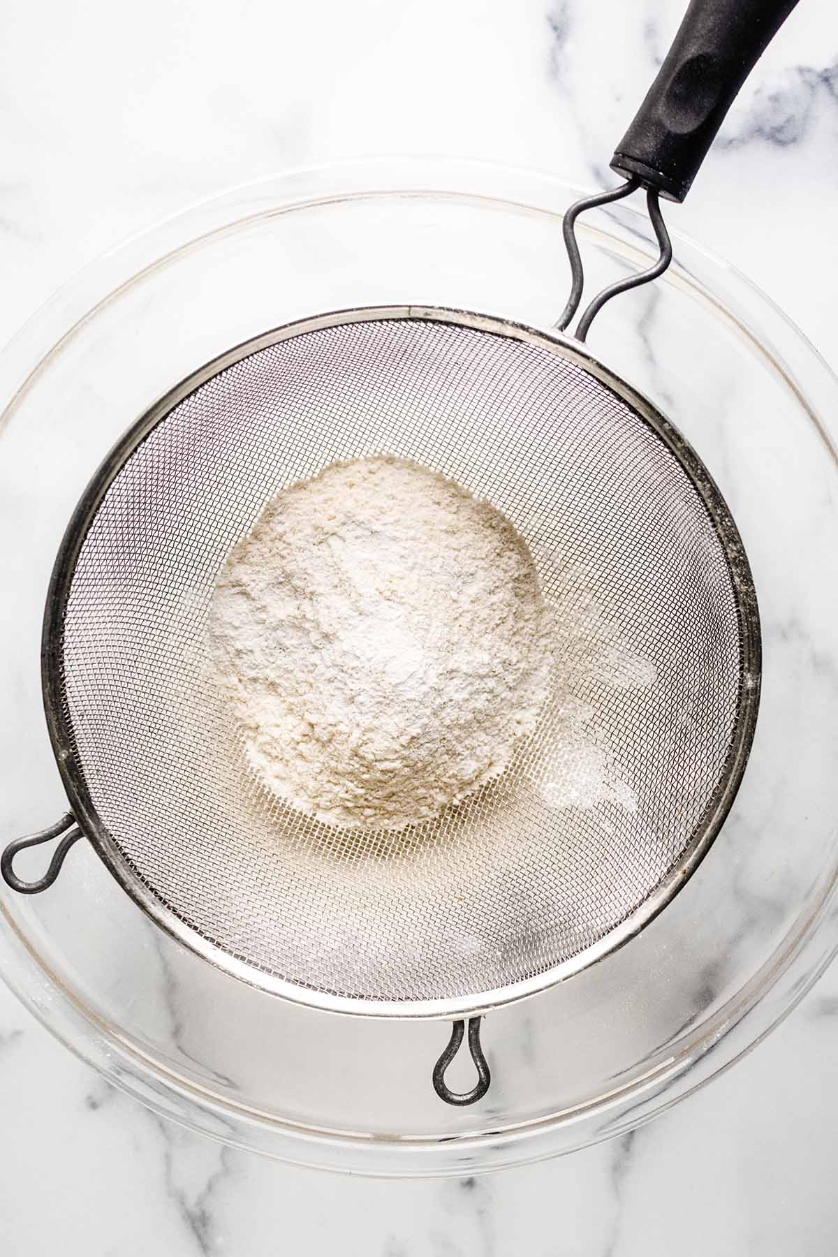 Overhead view of dry ingredients in a mesh strainer over a large glass bowl.