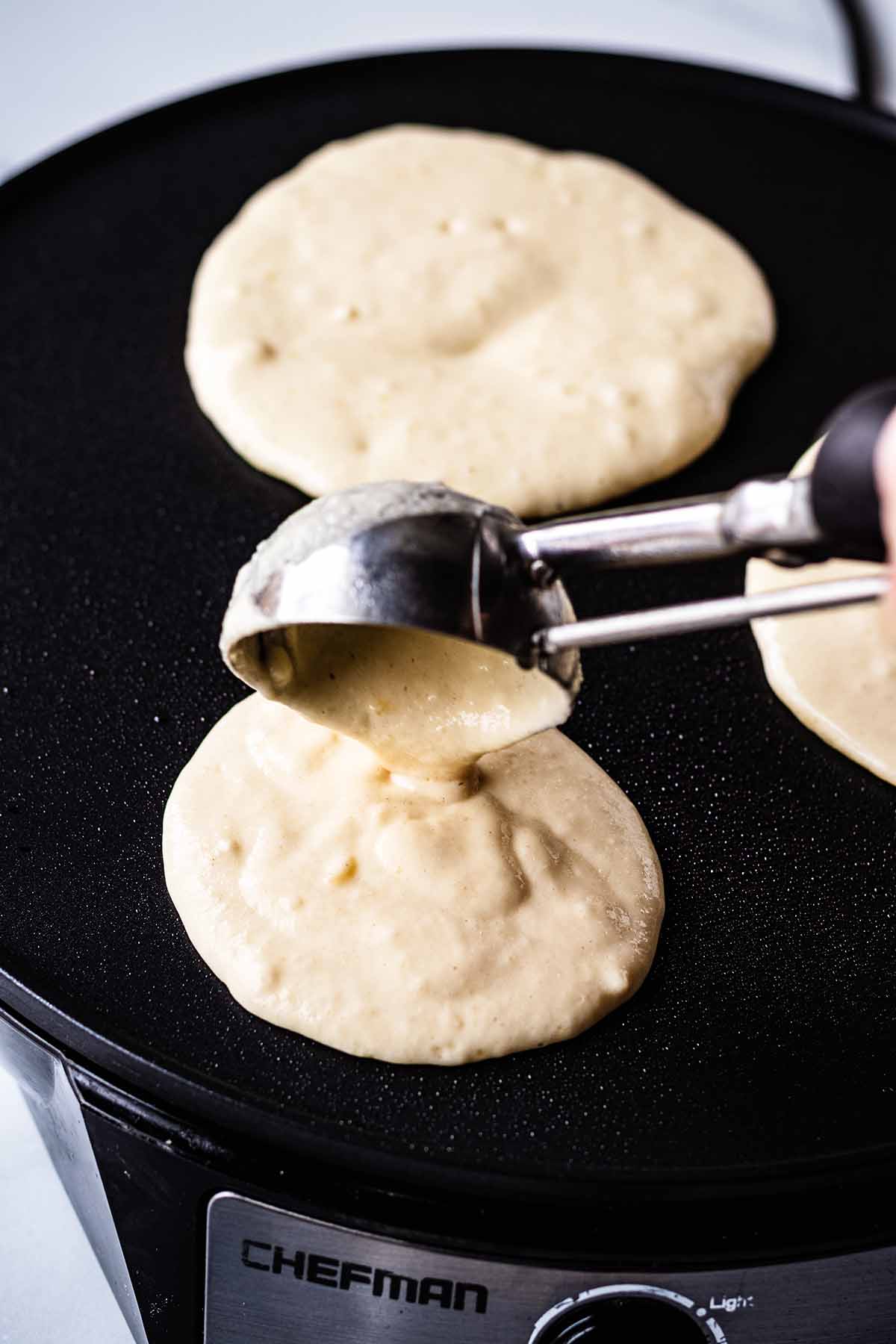 Pancake batter being poured onto a griddle with an ice cream scoop.