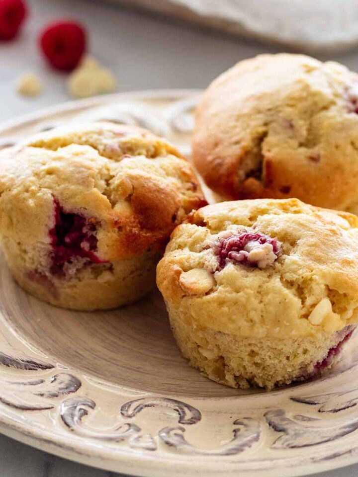 Close up of three muffins on a white plate. A bowl of muffins, raspberries, and white chocolate chips are in the background.