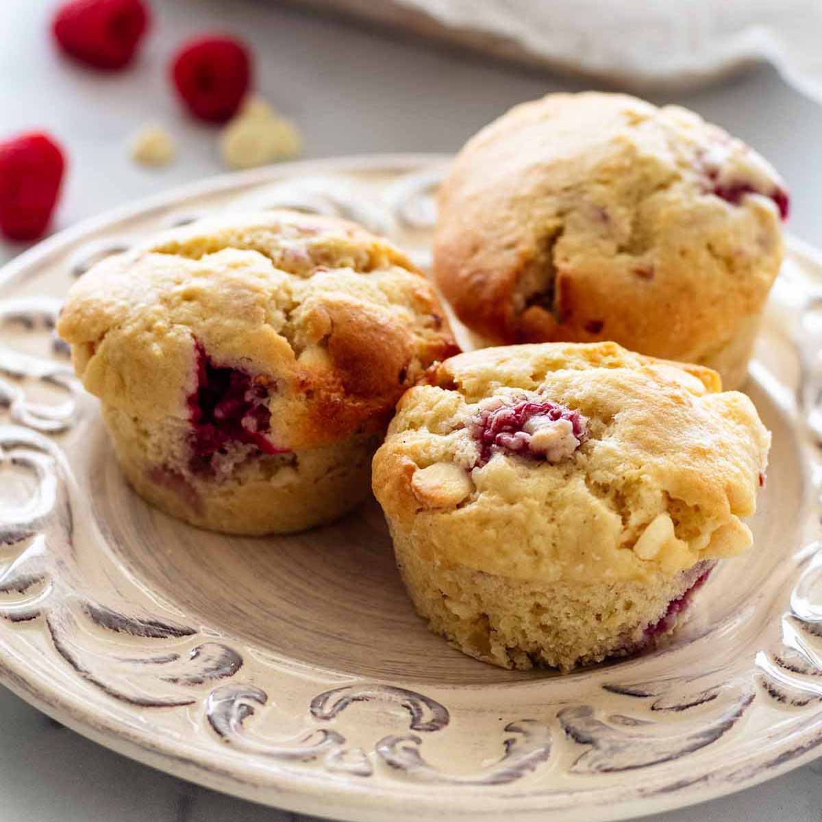 Close up of three muffins on a white plate. A bowl of muffins, raspberries, and white chocolate chips are in the background.