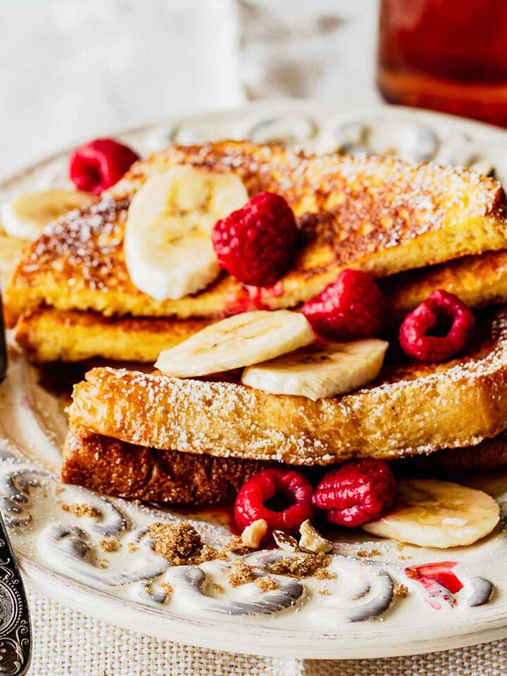 Stack of brioche French toast topped with sliced bananas and raspberries on a white plate with a fork.