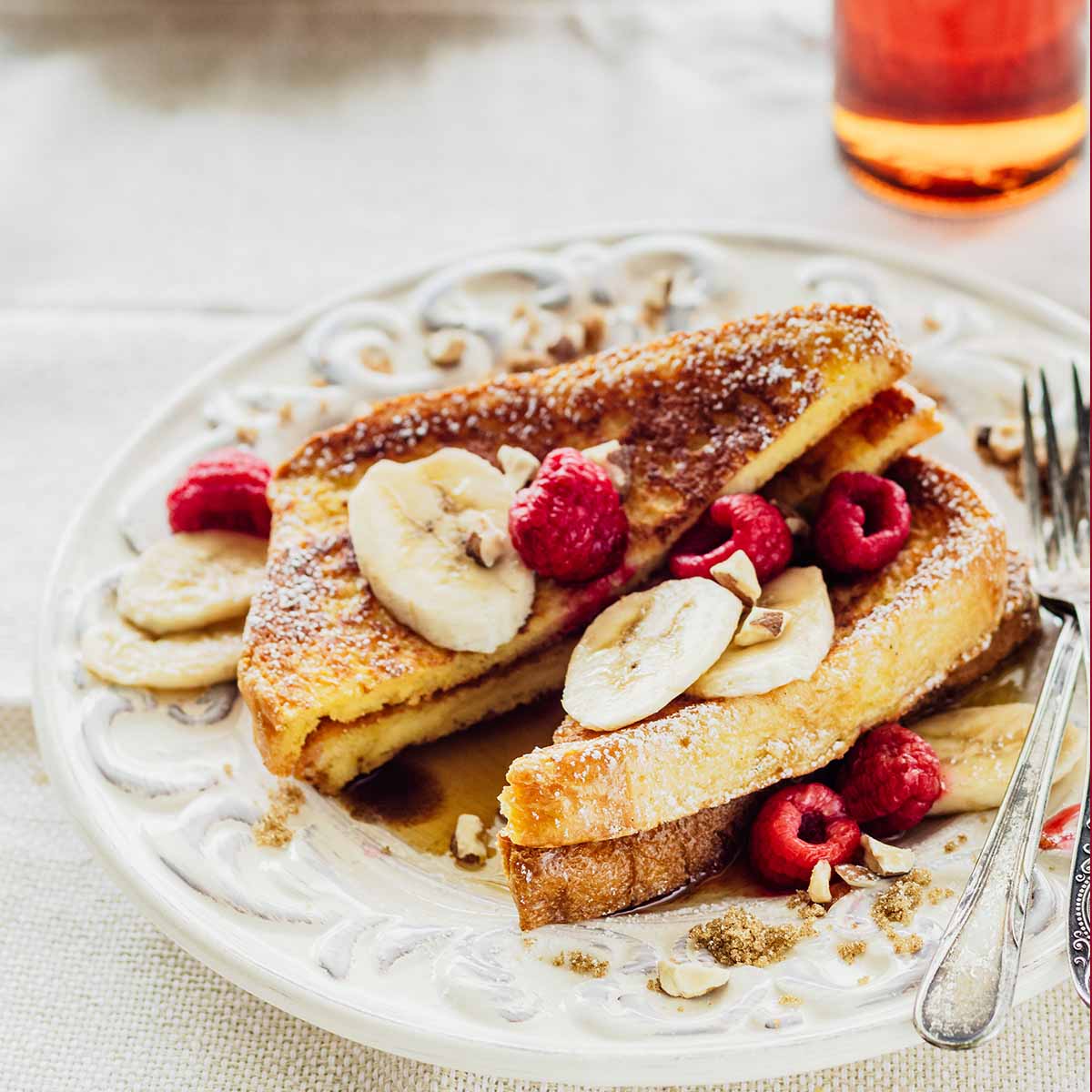 Stack of brioche French toast topped with sliced bananas and raspberries on a white plate with a fork.