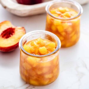 Two small jars of peach compote on a marble countertop with half of a fresh peach off to the side.