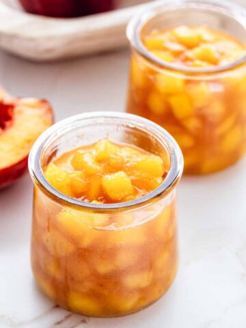 Two small jars of peach compote on a marble countertop with half of a fresh peach off to the side.