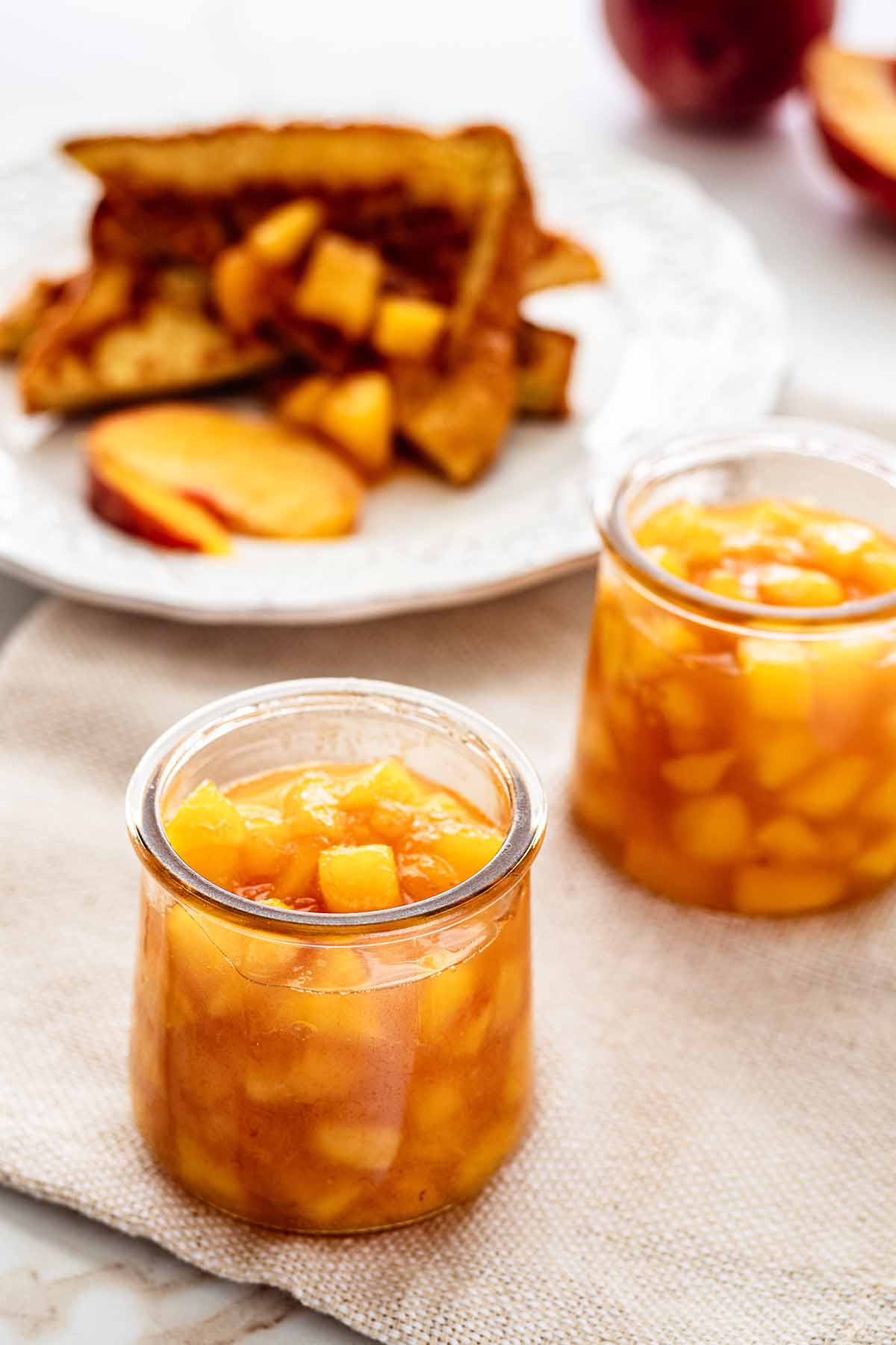 Two small jars of compote on a beige napkin with a plate of French toast in the background.