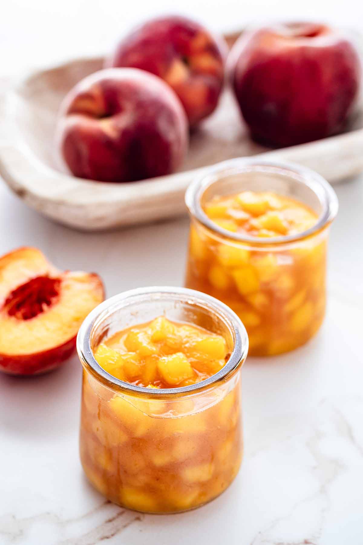 Two small jars of peach compote on a marble countertop with half of a fresh peach off to the side.