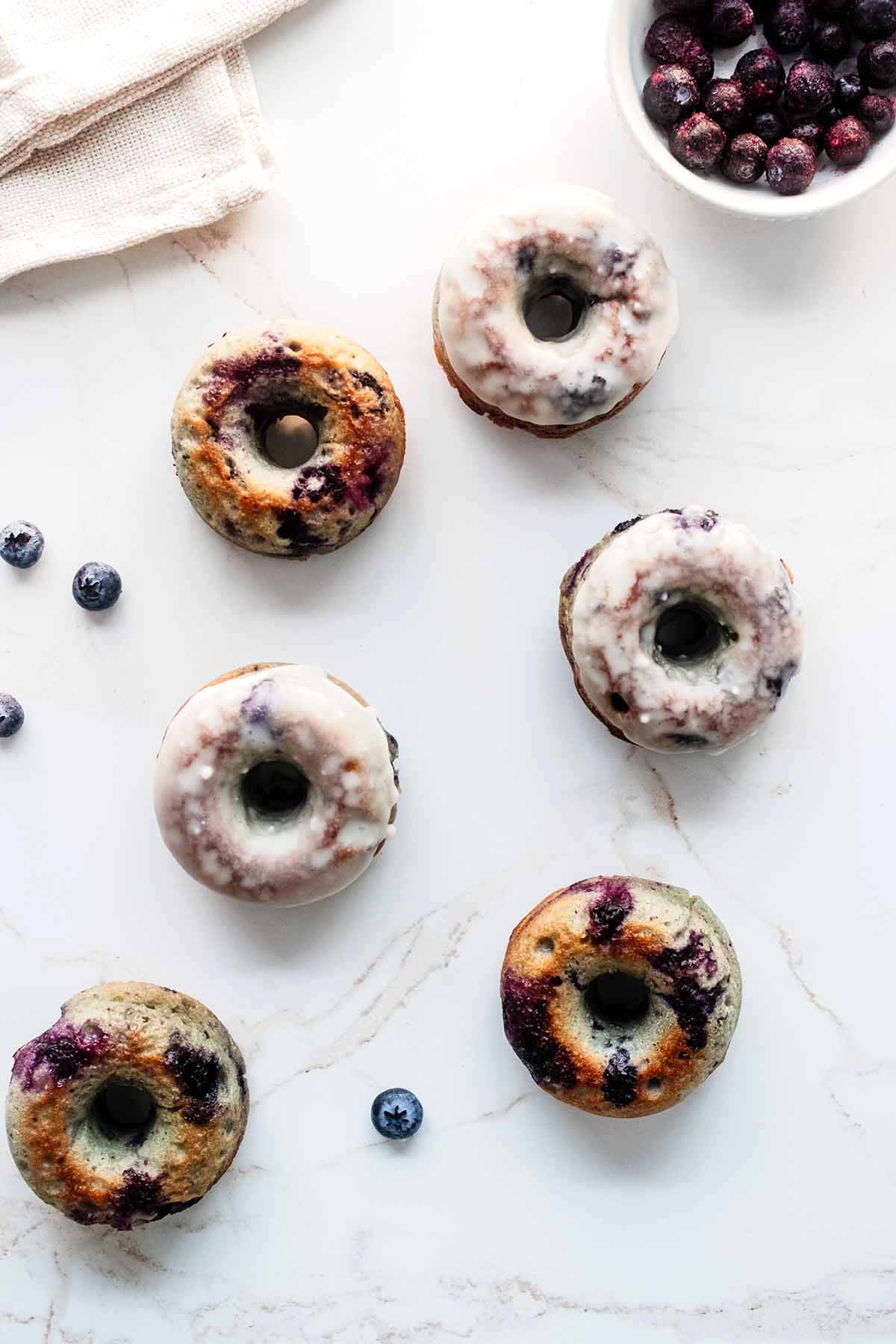 Overhead view of blueberry donuts on a marble surface with a bowl of blueberries and a white napkin