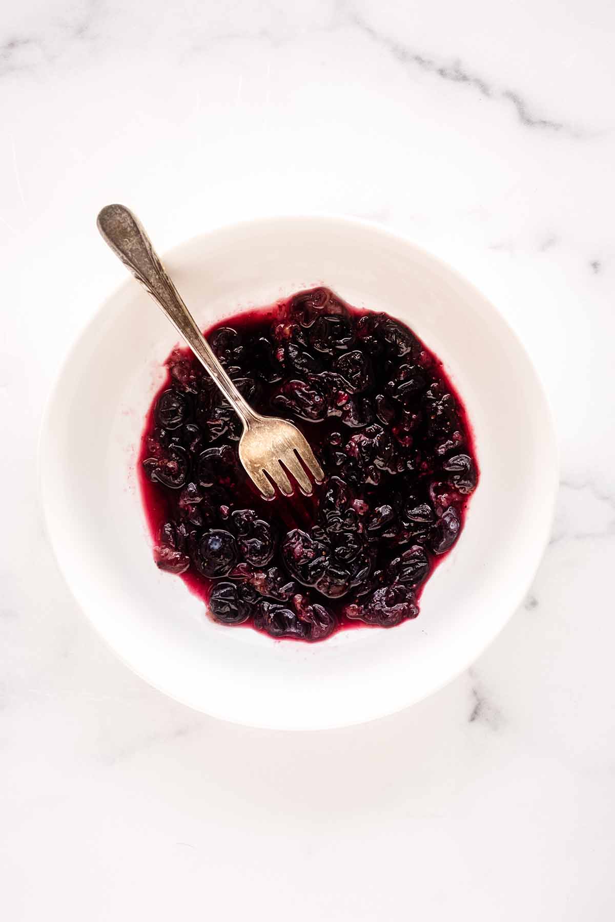 Overhead view of blueberries being crushed in a white bowl with a fork