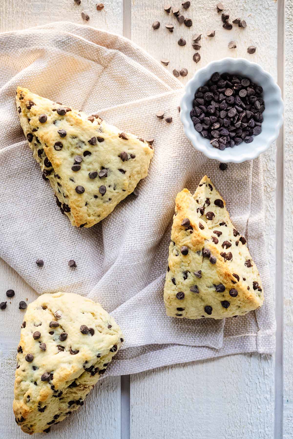 Overhead view of 3 chocolate chip scones on a cream napkin on a white wood background with a small white bowl filled with chocolate chips