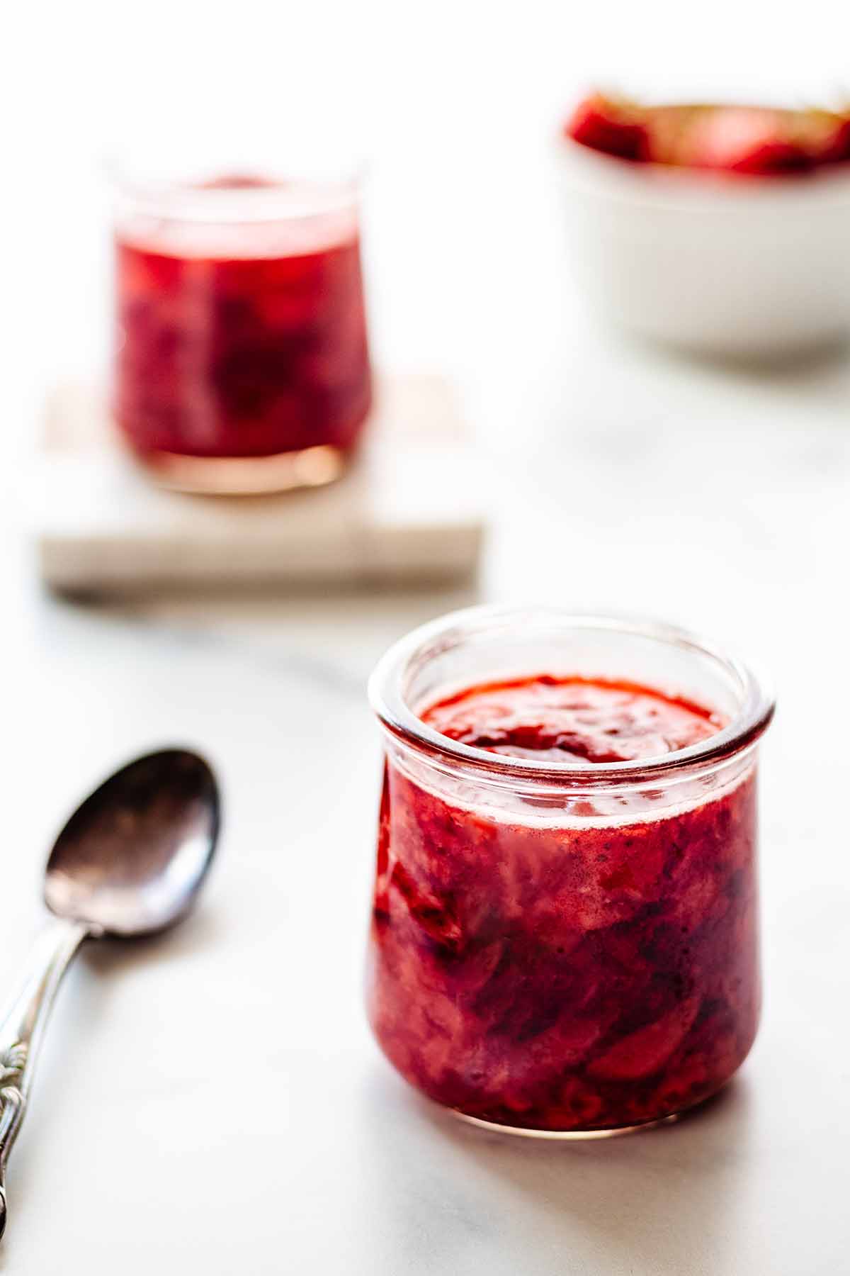 Two small jars of compote on a marble background with a spoon and small bowl of strawberries