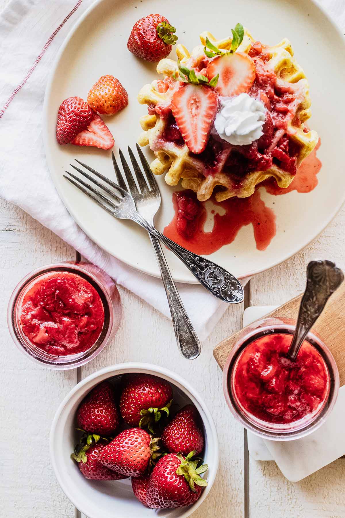Overhead view of 2 jars of strawberry compote, a bowl of strawberries, and compote-covered waffles on a white plate with a fork