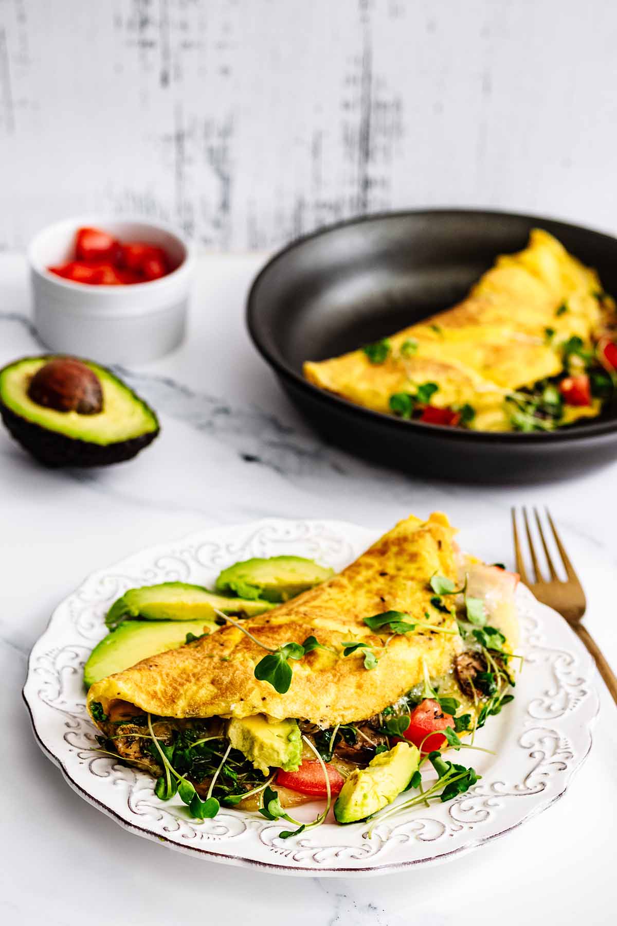 Loaded California omelette on a white plate with avocado slices and a gold fork. A second omelette is in a skillet int the background.