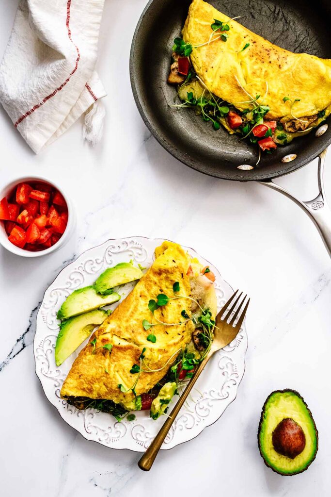 Overhead view of California omelette on a white plate with a gold fork and sliced avocado next to a second omelette in a skillet