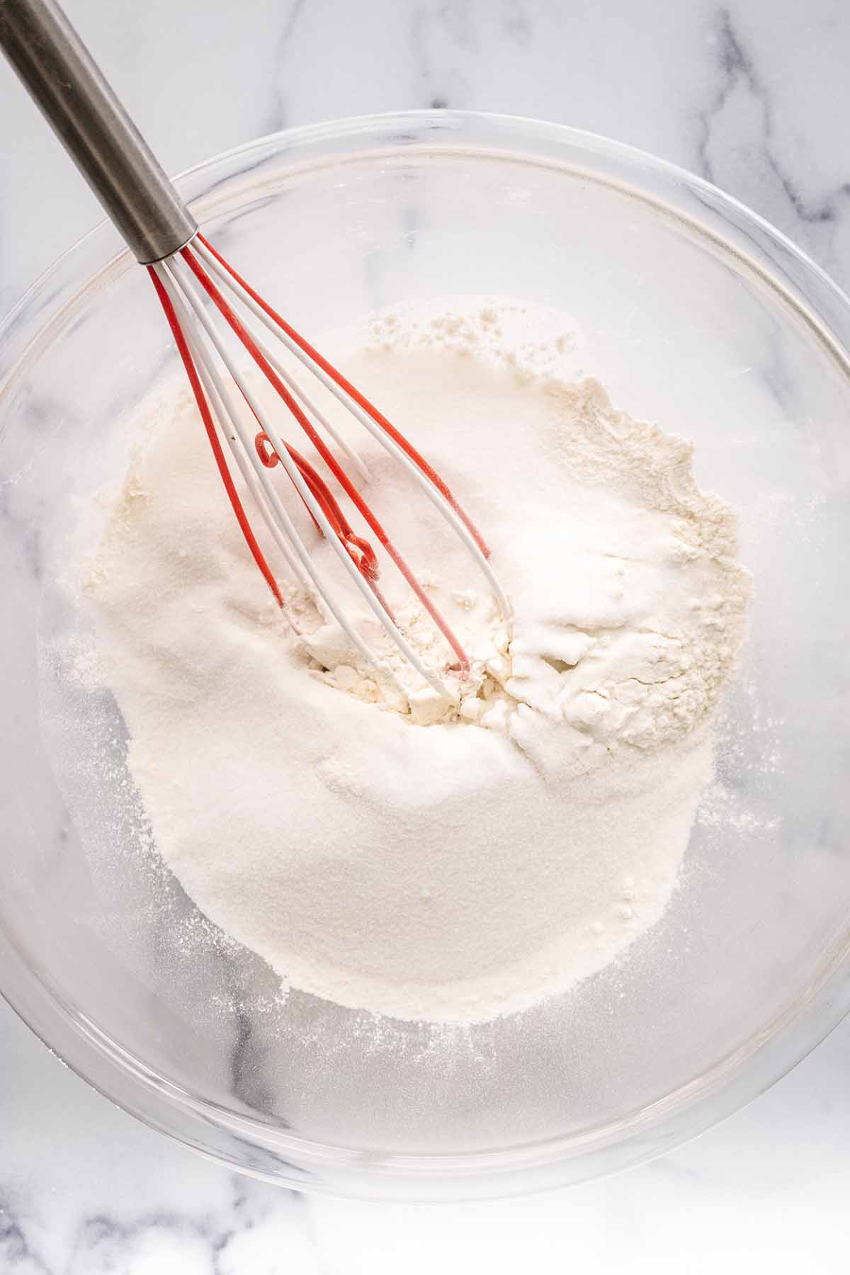 Overhead view of dry ingredients in a large glass bowl with a red and white silicone whisk