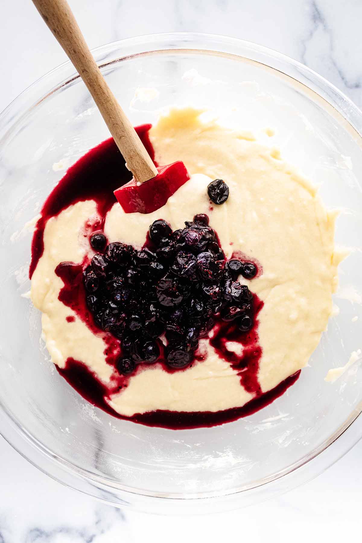 Overhead view of crushed blueberries in muffin batter with a red spatula in a glass bowl