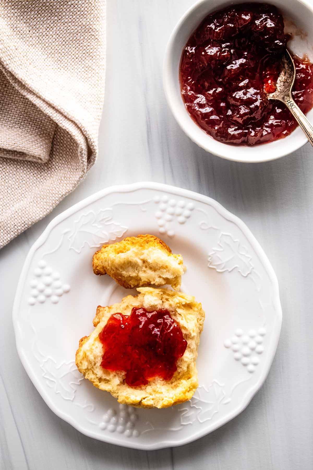 Half a biscuit topped with strawberry jam on a white plate with a small bowl of strawberry jam.