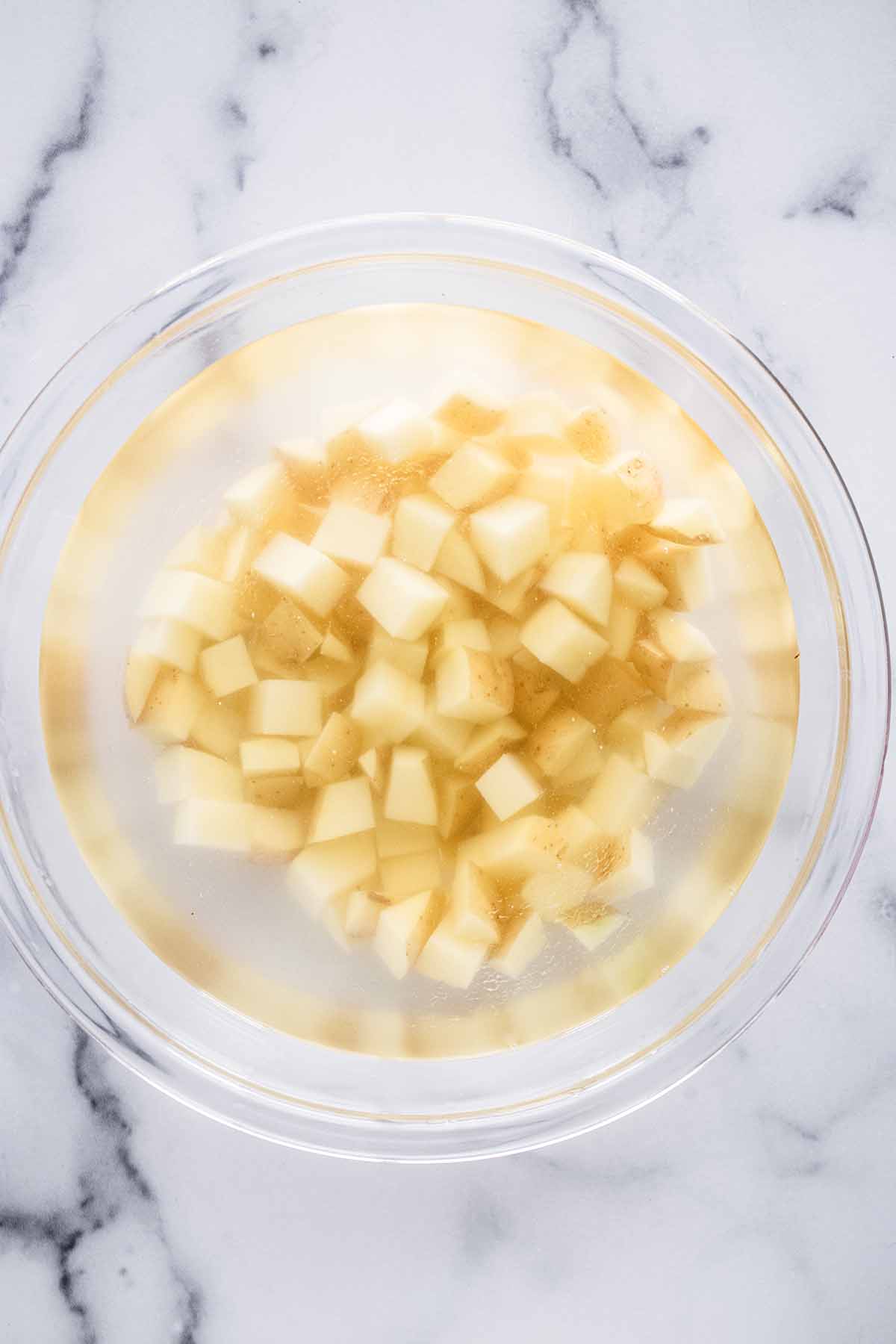 Overhead view of cubed potatoes soaking in water in a glass bowl.