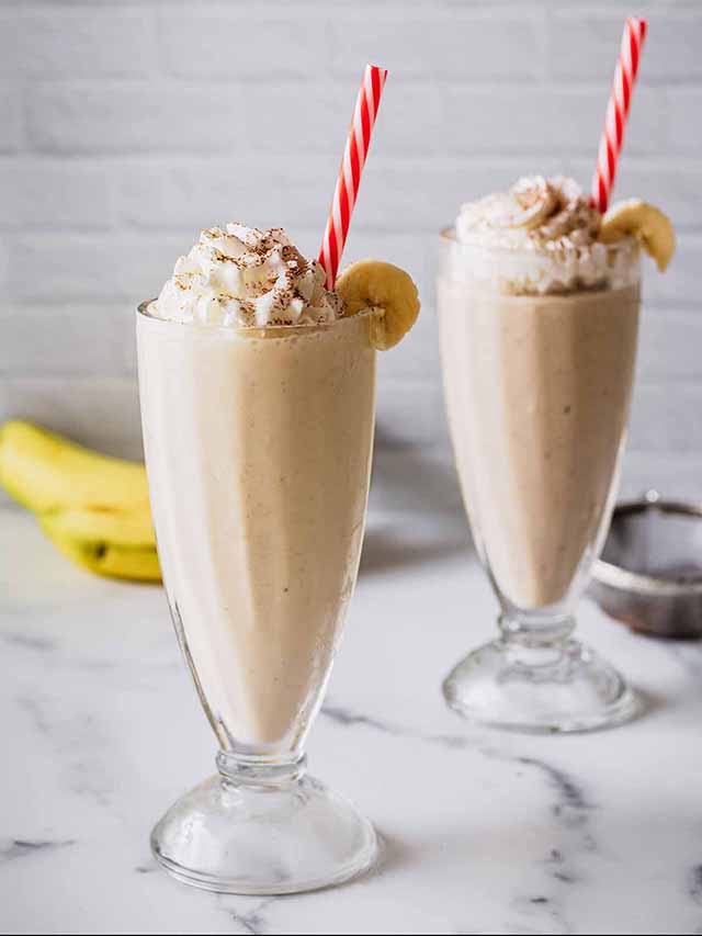 Two banana milkshakes in tall glasses with whipped cream and straws.