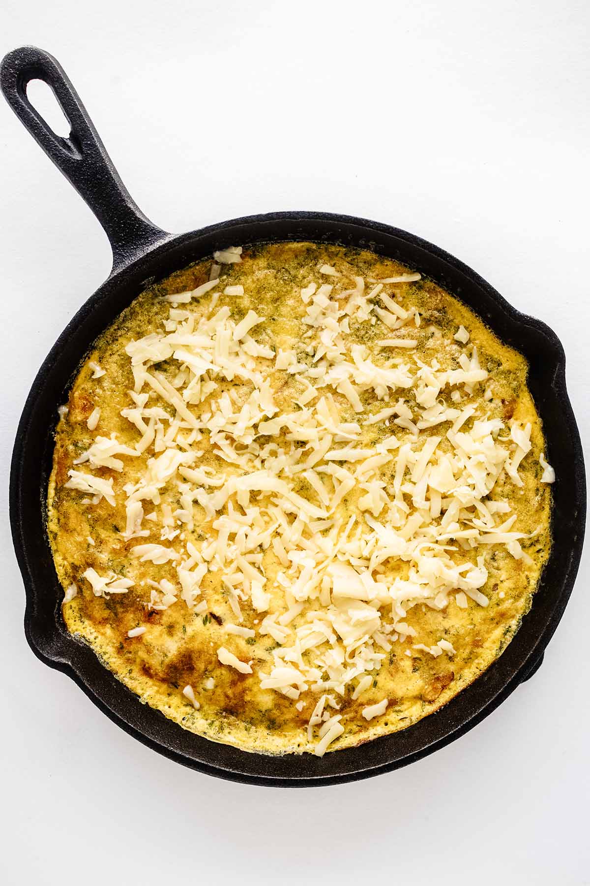 Overhead view of partially cooked frittata topped with grated cheese in a cast iron skillet.