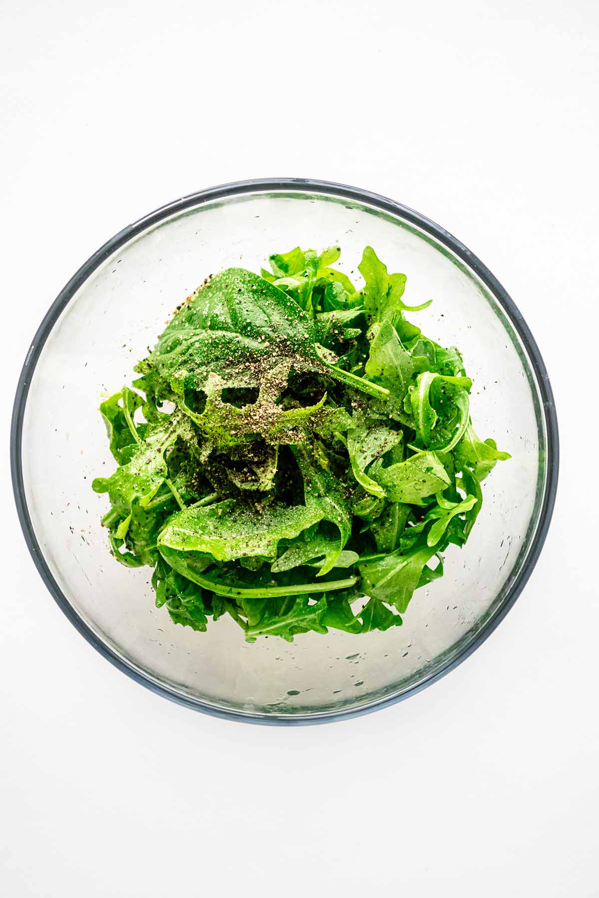 Overhead view of arugula, oil, salt, and pepper in a small glass bowl.