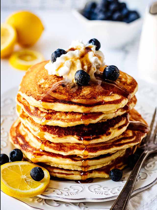 Stack of lemon blueberry pancakes topped with whipped cream and fresh blueberries on a white plate.