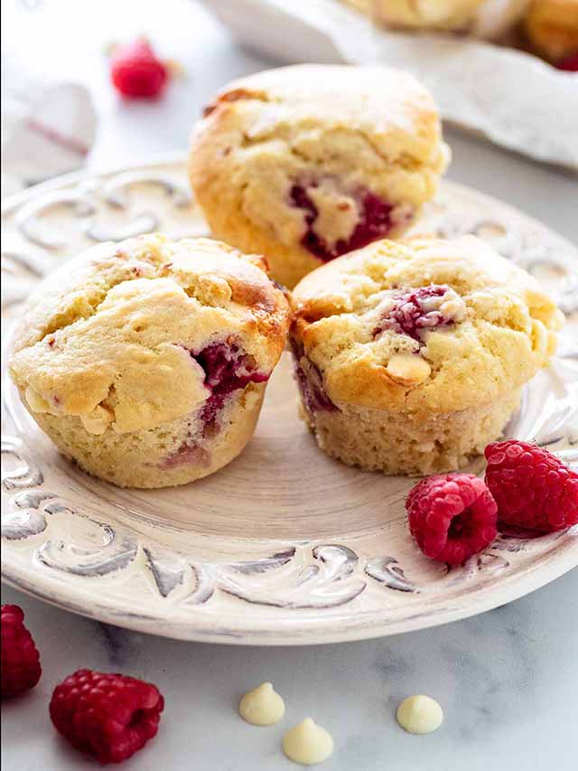 Three raspberry and white chocolate muffins on a white plate with fresh raspberries