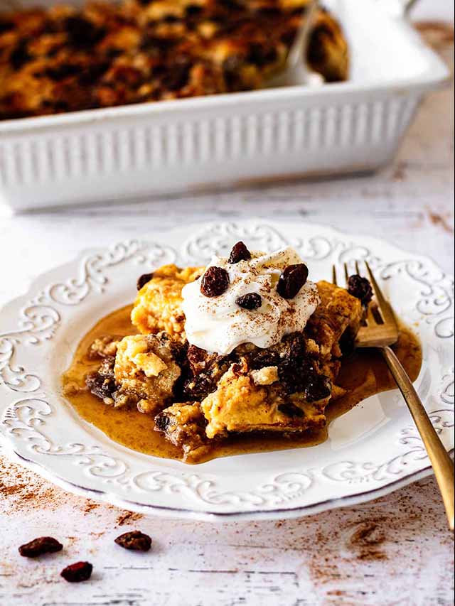 Serving of raisin bread pudding topped with whipped cream and raisins on a white plate with a gold fork.