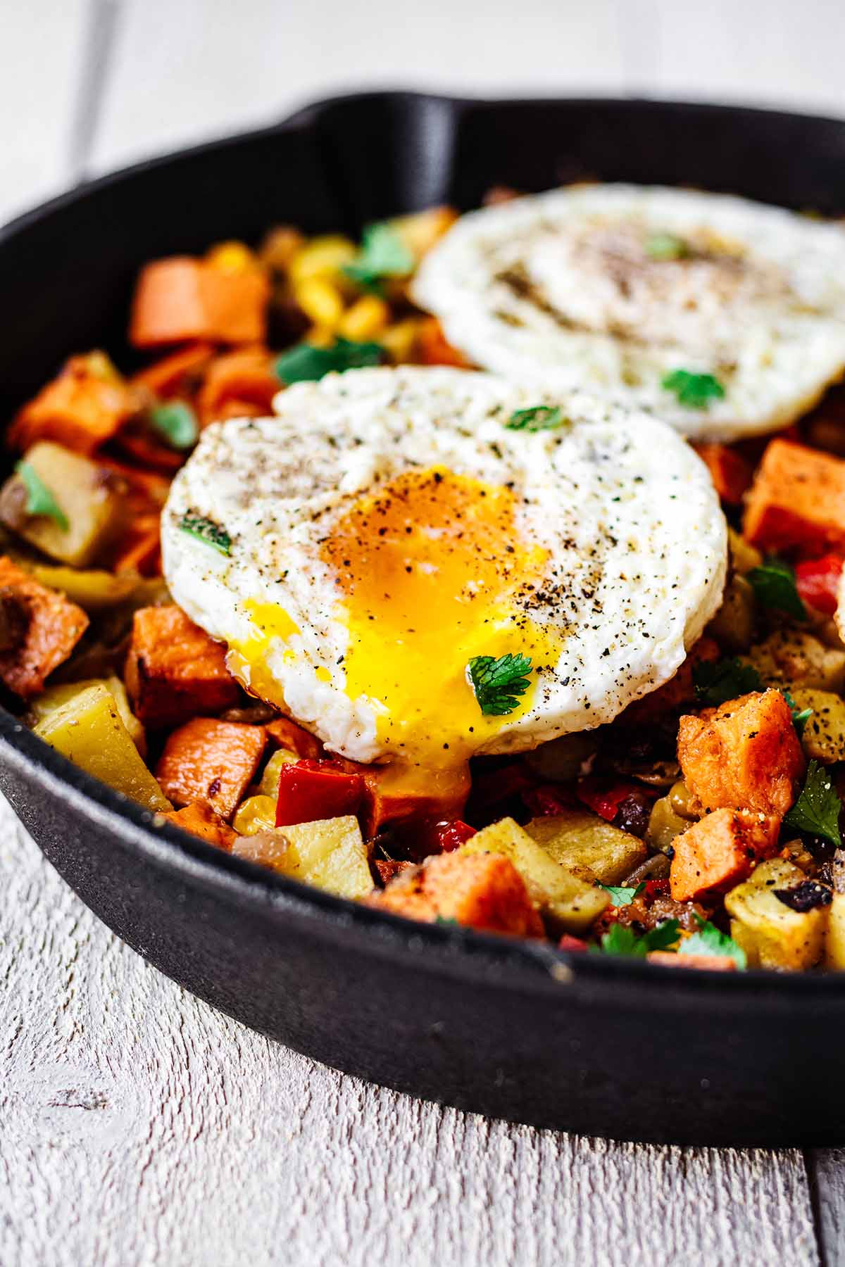 Close up of sweet potato breakfast hash topped with two fried eggs with runny yolk in a cast iron skillet.