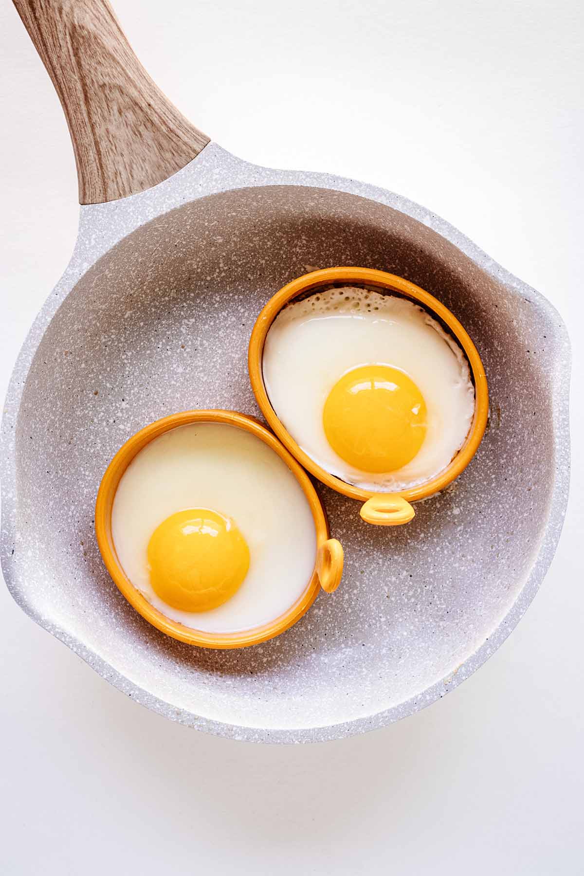 Two eggs in egg rings cooking in a small white skillet with a light wooden handle.