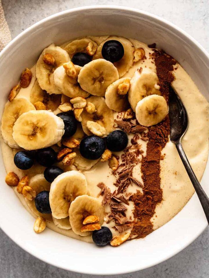 Banana smoothie bowl topped with chopped peanuts, fresh blueberries, chocolate shavings, and a line of ground cinnamon in a white bowl with a spoon.