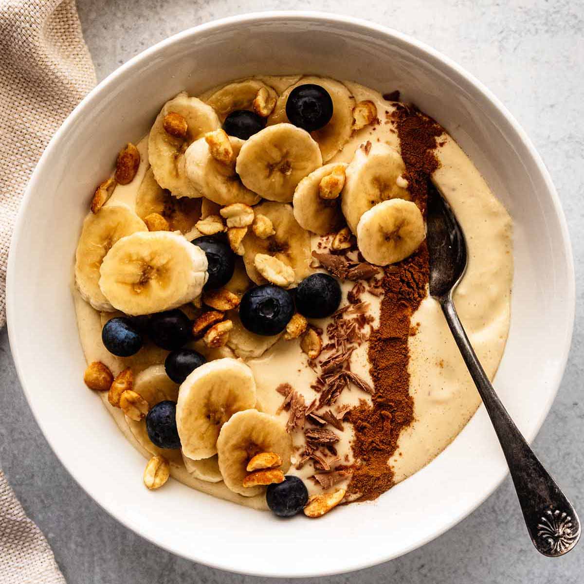 Banana smoothie bowl topped with chopped peanuts, fresh blueberries, chocolate shavings, and a line of ground cinnamon in a white bowl with a spoon.